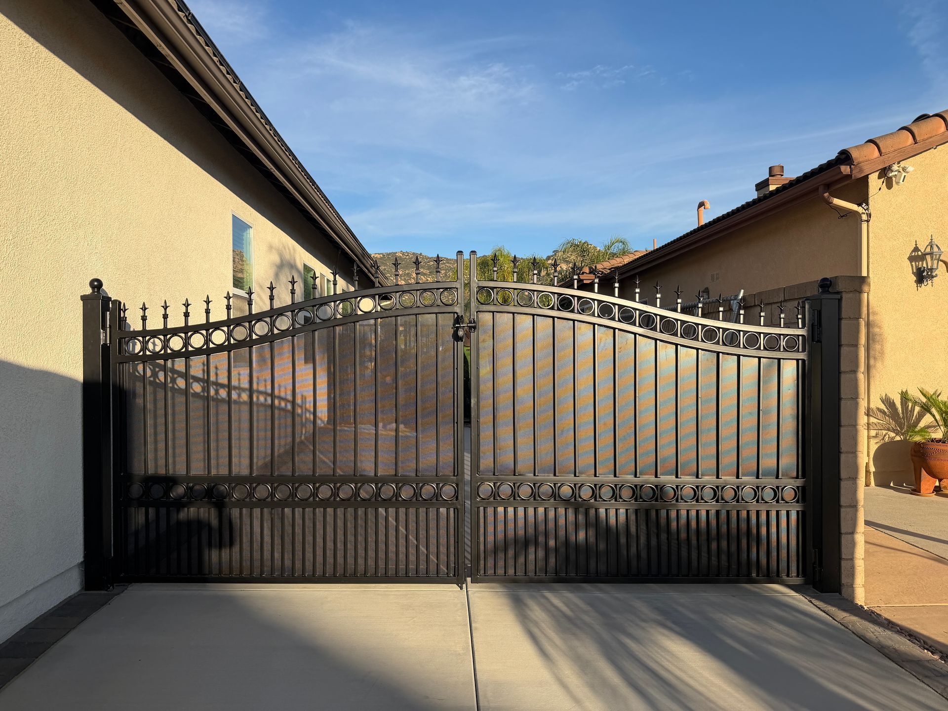 Black wrought-iron driveway gate between yellow stucco walls under a clear blue sky