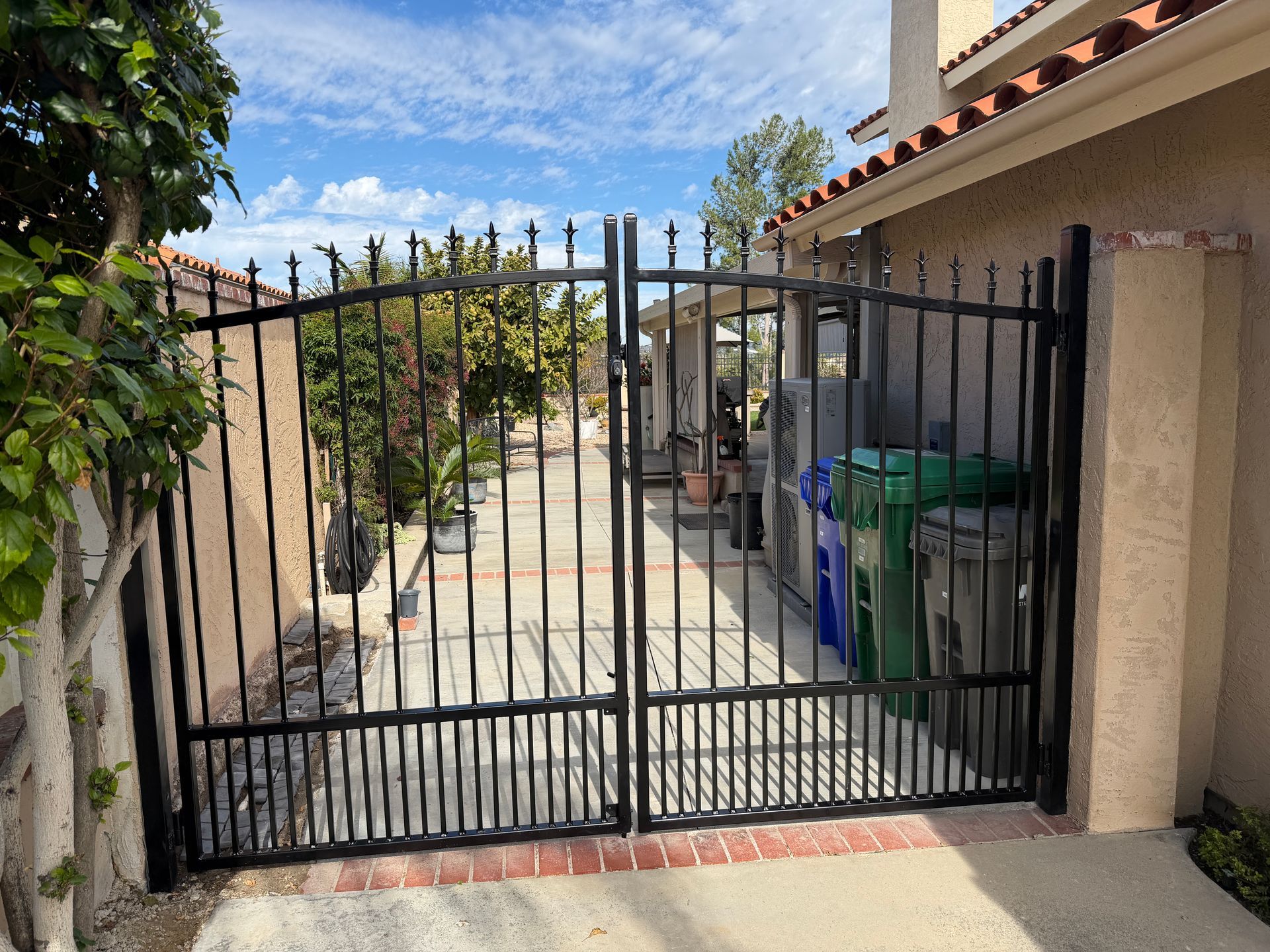 Black metal gate at a residential driveway with trash bins and a stucco wall under a blue sky