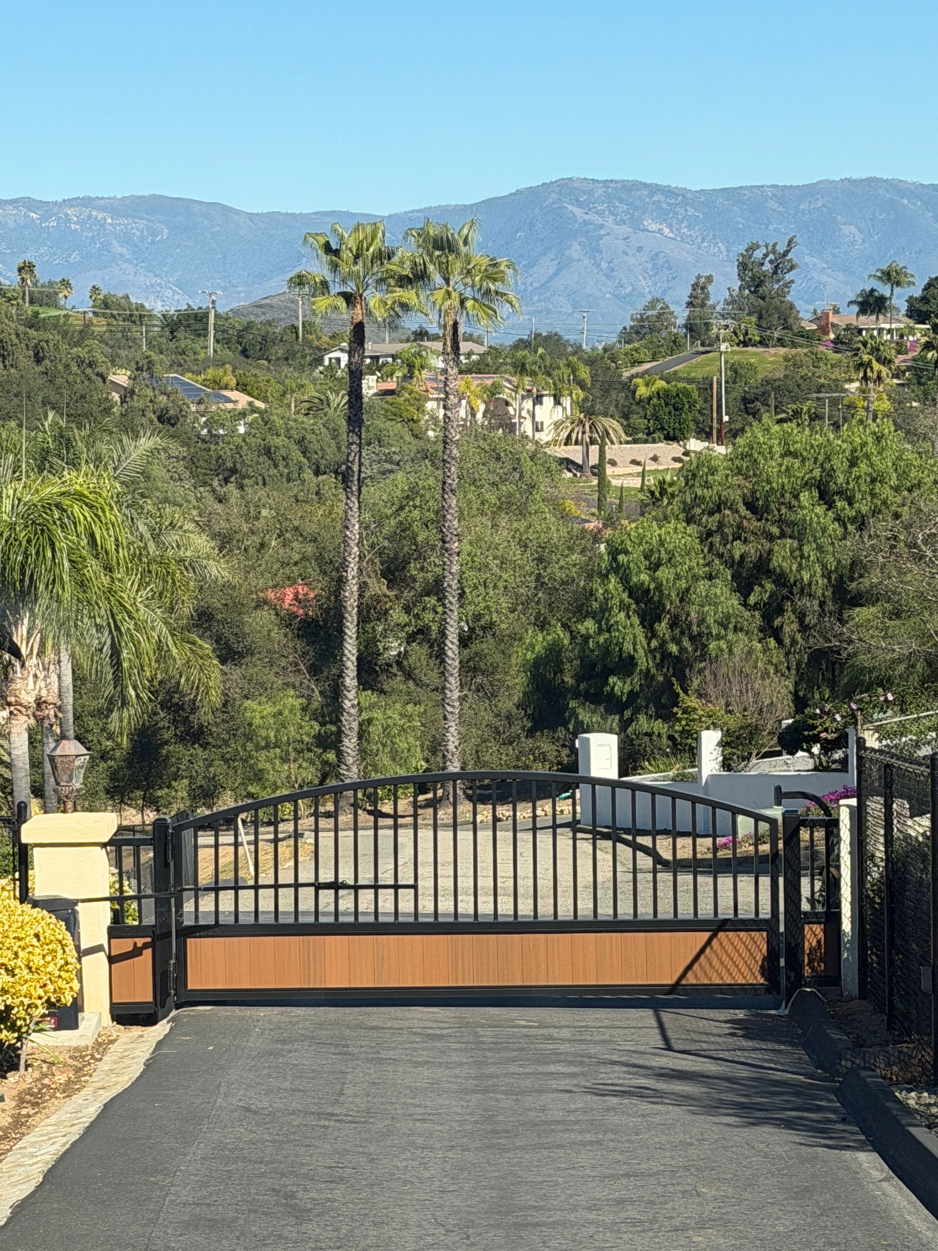 Closed black gate at the end of a driveway, with trees and mountains in the background.