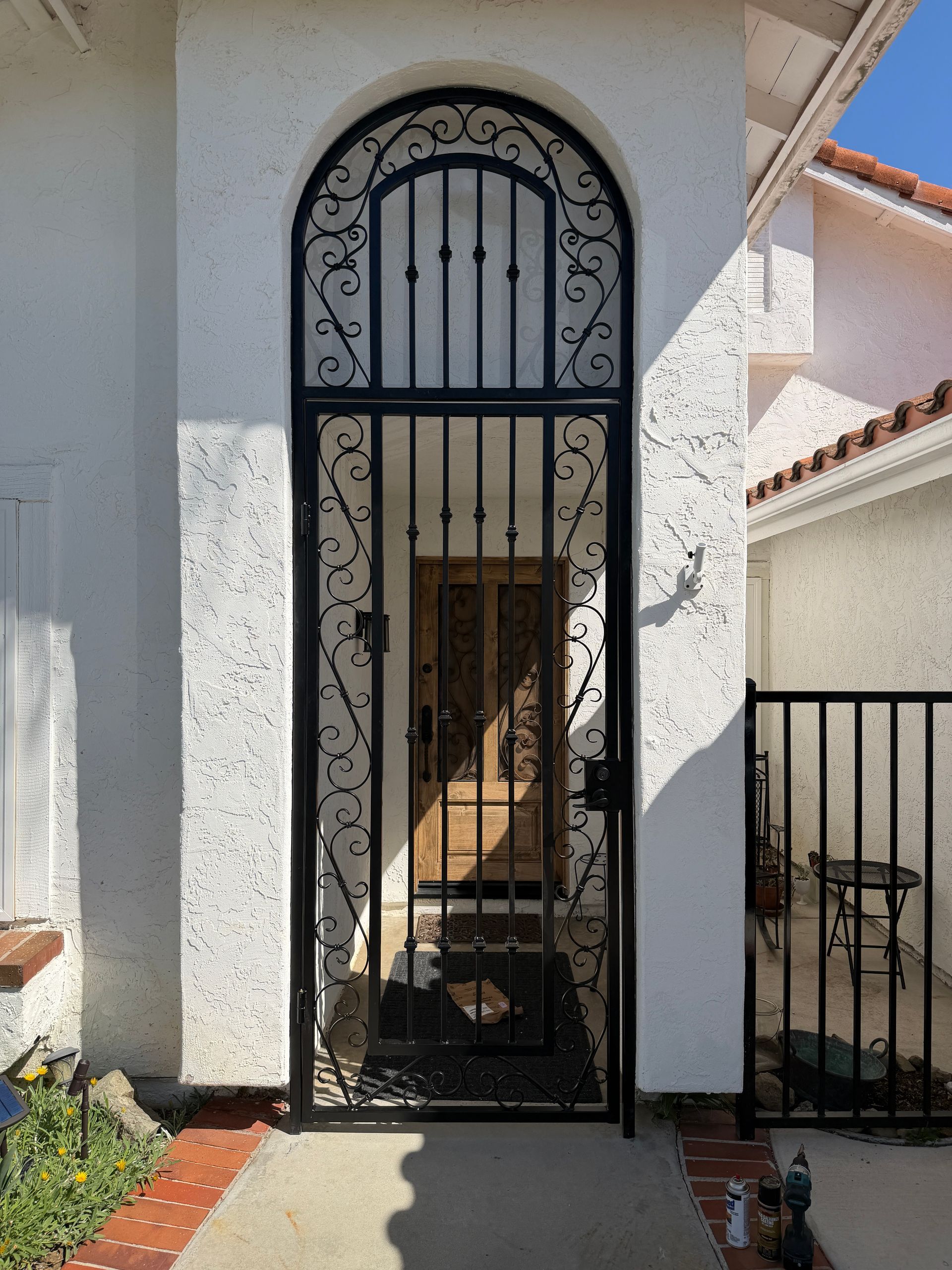 Black wrought-iron security gate at a house entrance, with arched top and stucco walls.