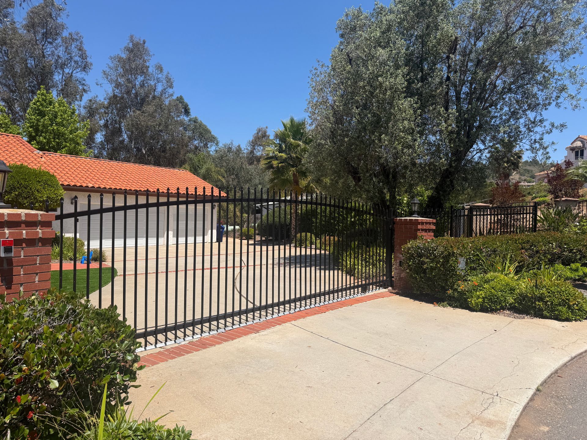 Black metal driveway gate beside a brick wall and trimmed hedge, with a concrete driveway and trees behind.