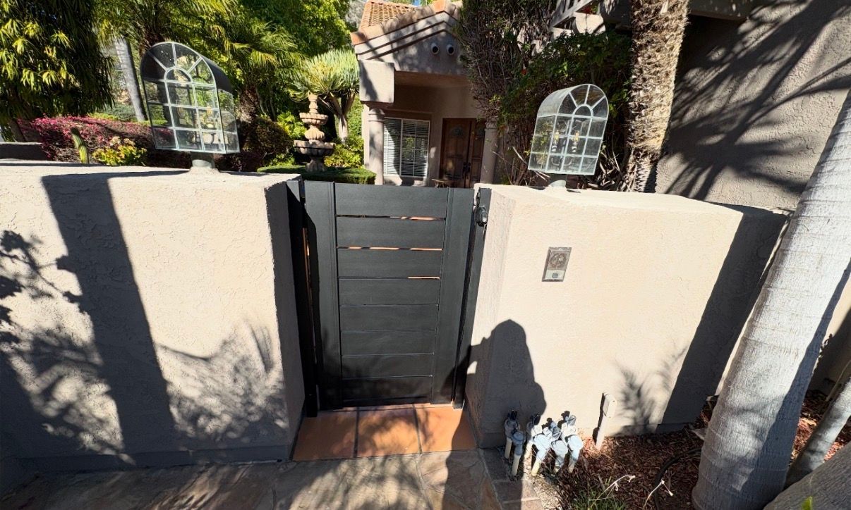 Front gate entrance to a house with cream walls, dark door, and sunlit garden shadows.