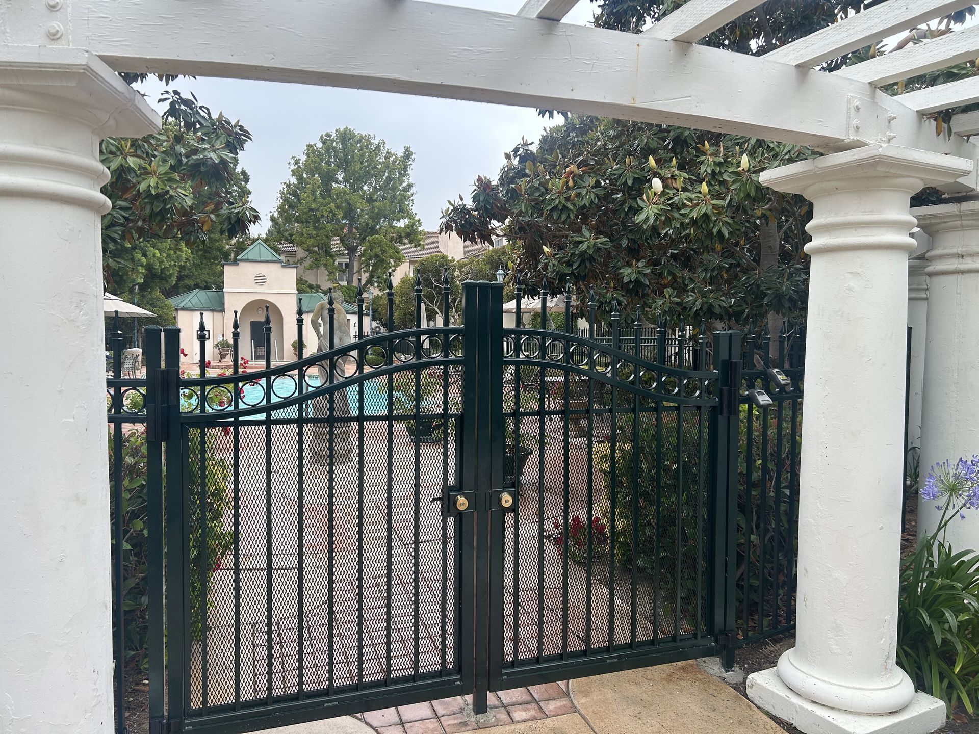 Black wrought-iron gate under white columns, opening to a tree-lined courtyard and pool area