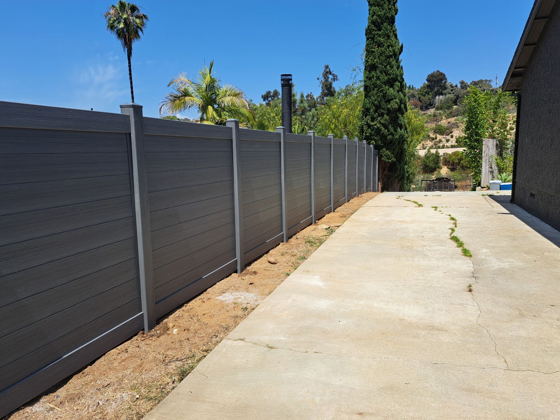 Long concrete driveway beside a tall gray fence, with palm trees and hillside in the background