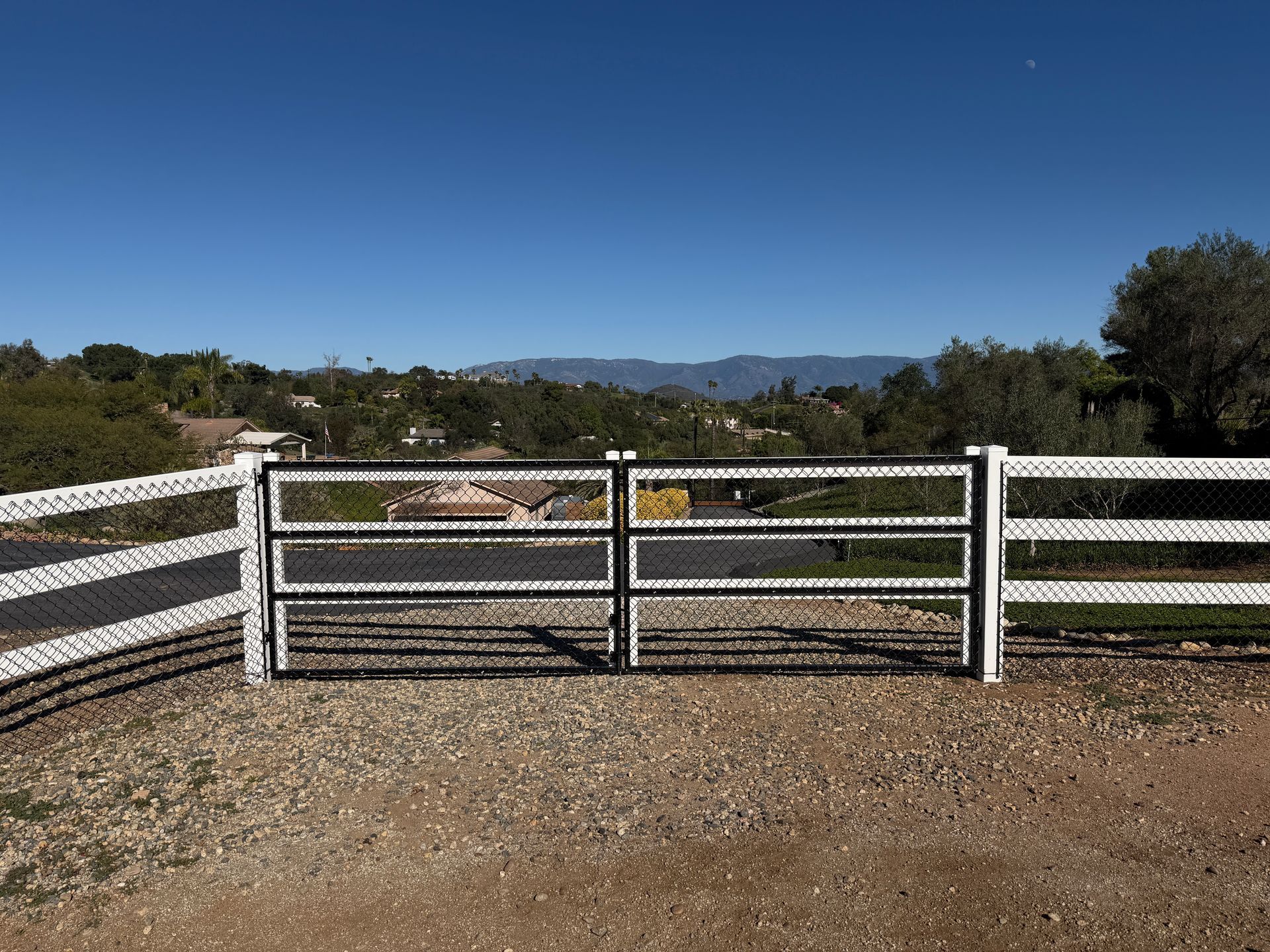 White ranch gate over gravel driveway with hills and blue sky in the background