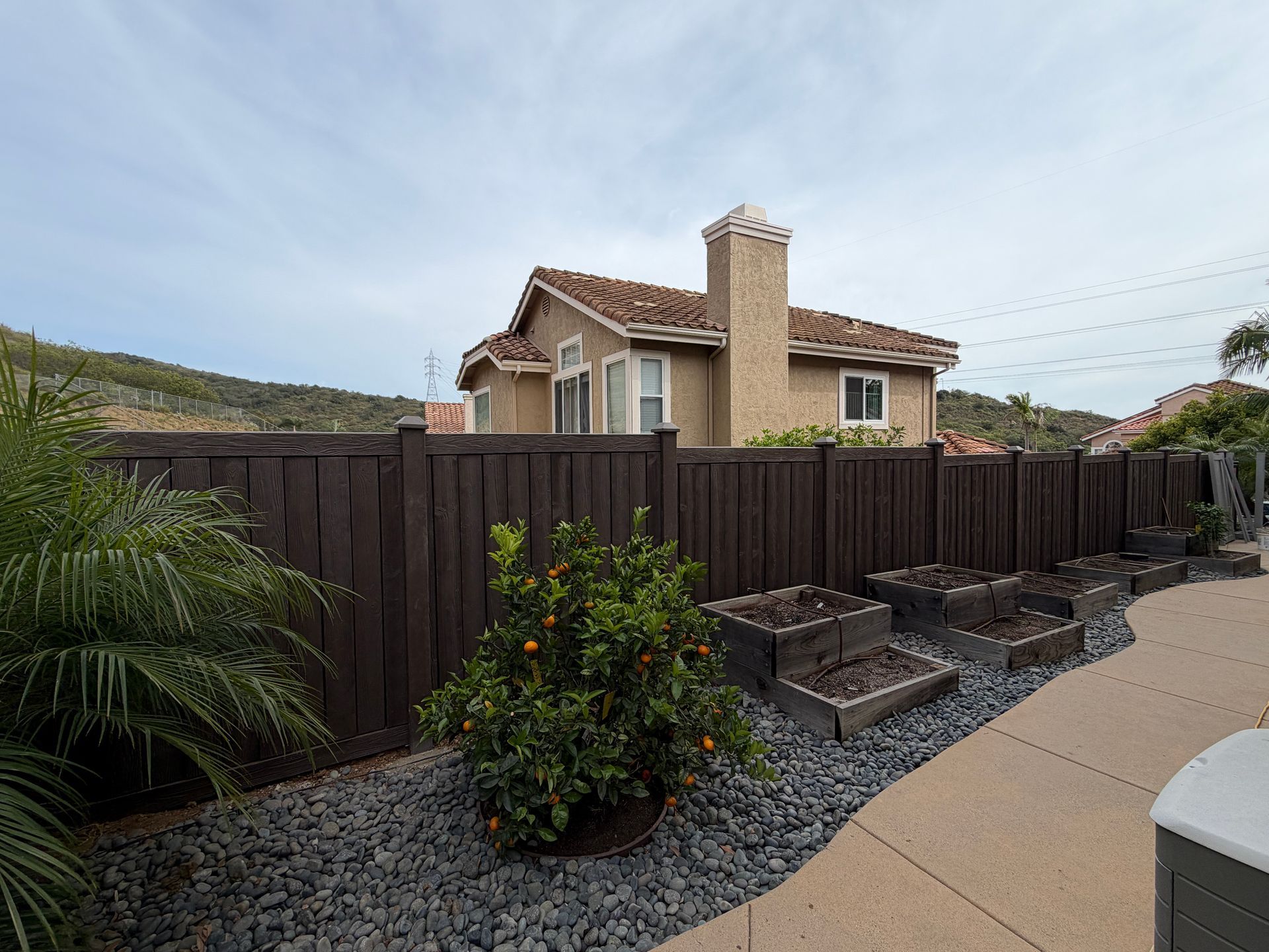 Suburban house behind a dark wooden fence with a gravel yard and a cloudy sky
