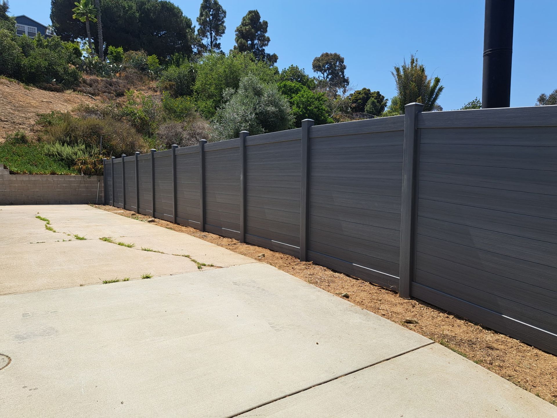 Long gray fence lining a sloped driveway beside dry landscaping and trees under a blue sky