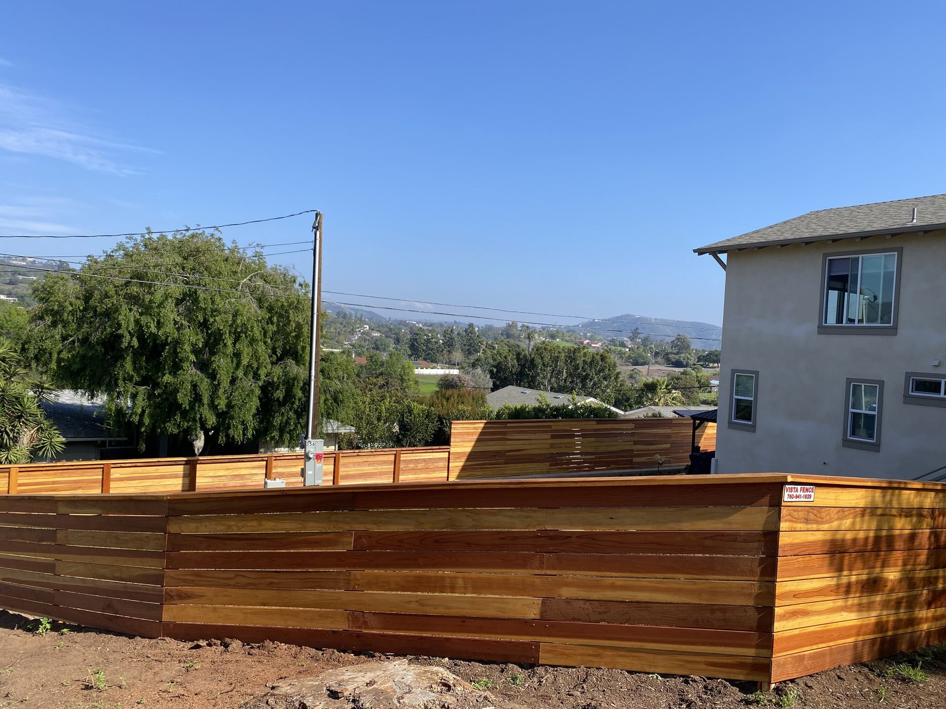 Wooden fenced backyard with trees, a house, and distant mountains under a clear blue sky