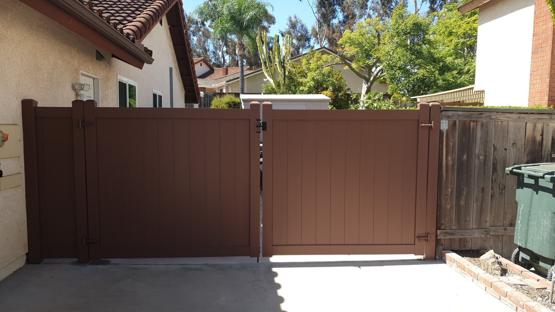 Brown wooden driveway gate between two stucco houses, with a sunny courtyard and trash bins on the right.