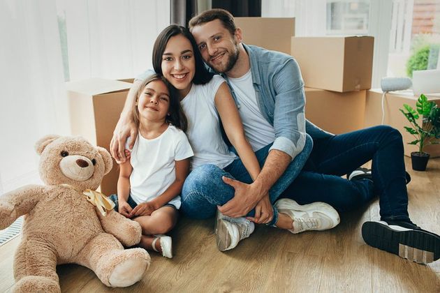 Family sitting on the floor in a bright living room with a teddy bear and moving boxes, smiling at the camera