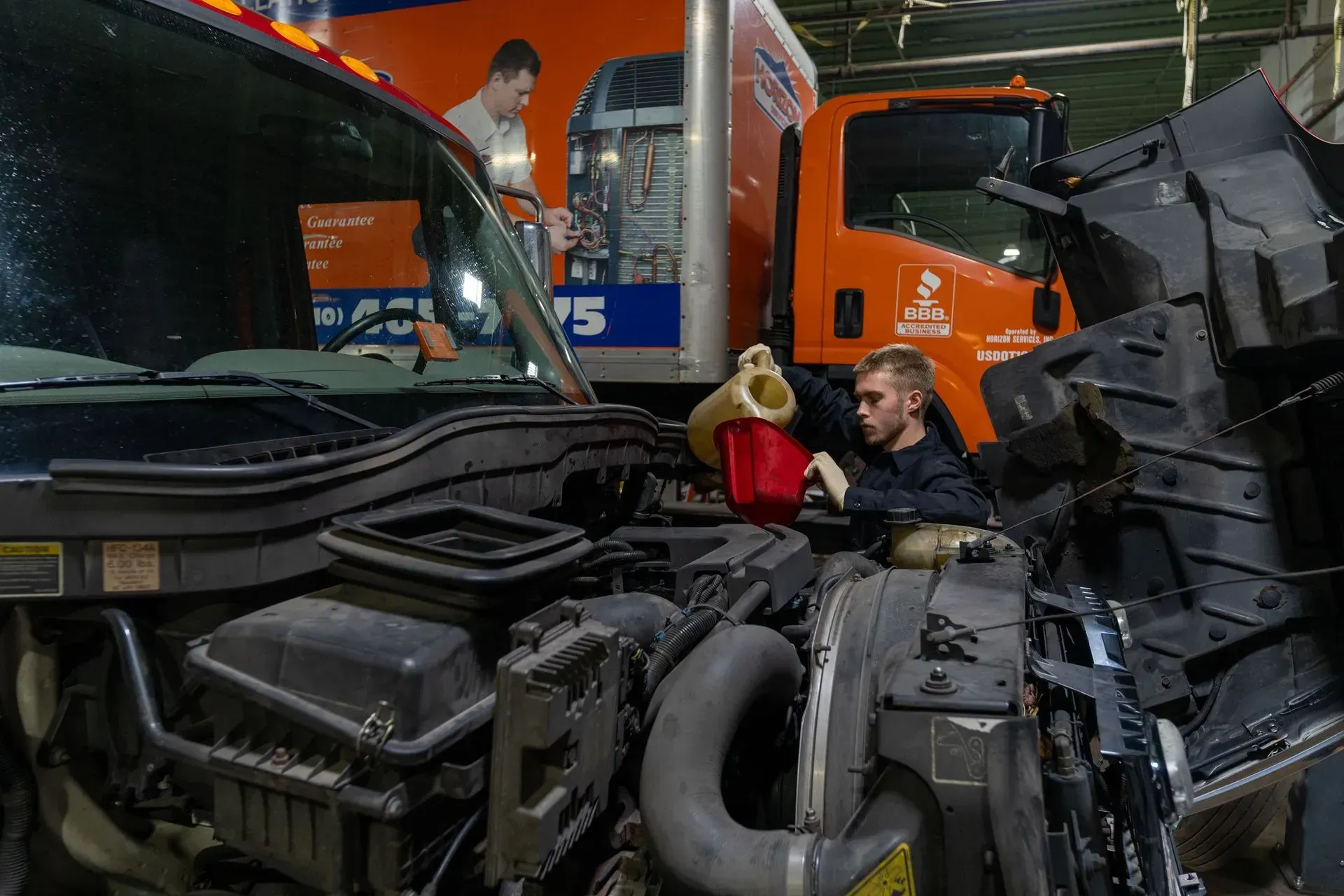 Mechanic pouring fluid into a truck engine in a garage. An orange truck and a poster are in the background.
