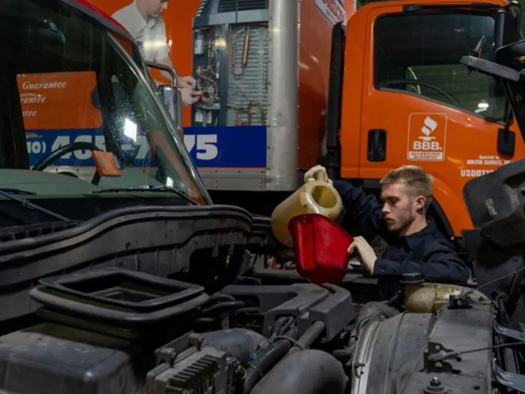 Mechanic pouring fluid into a car engine, orange truck in the background.