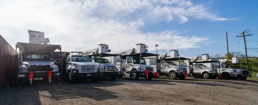 Several white utility trucks parked on gravel under a partly cloudy sky.