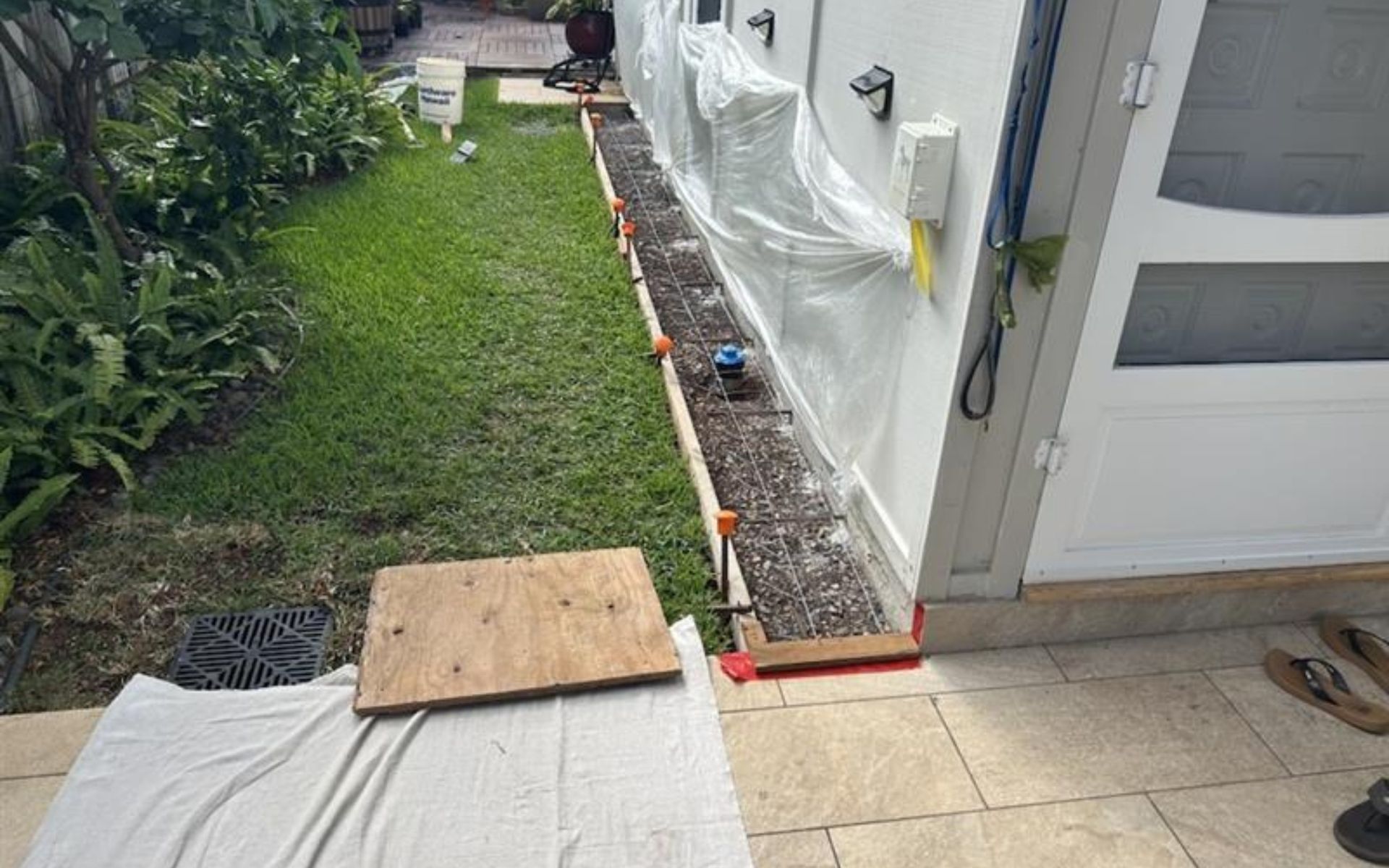 Narrow side yard beside a house, with a plywood ramp over a step and red-edged walkway under construction