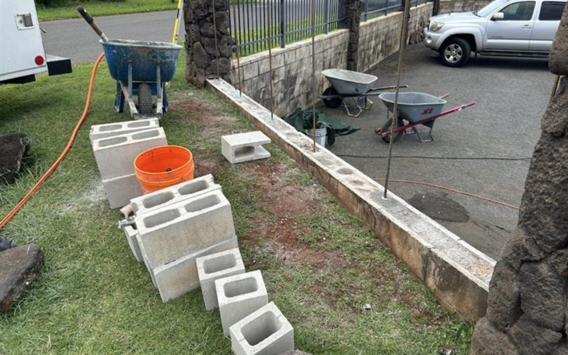 Construction materials and wheelbarrows beside a low stone wall in a yard, with cinder blocks and an orange bucket.