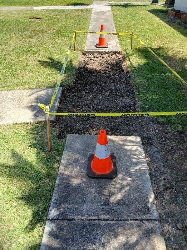 Broken sidewalk under repair, marked with caution tape and orange traffic cones on a grassy residential path