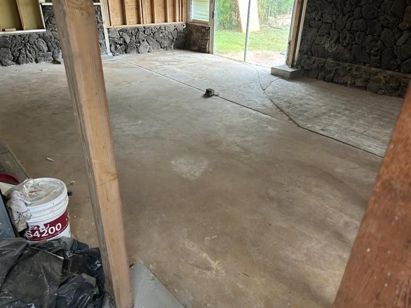 Covered concrete floor under a rustic open-air structure with stone walls and wooden posts; bucket at left