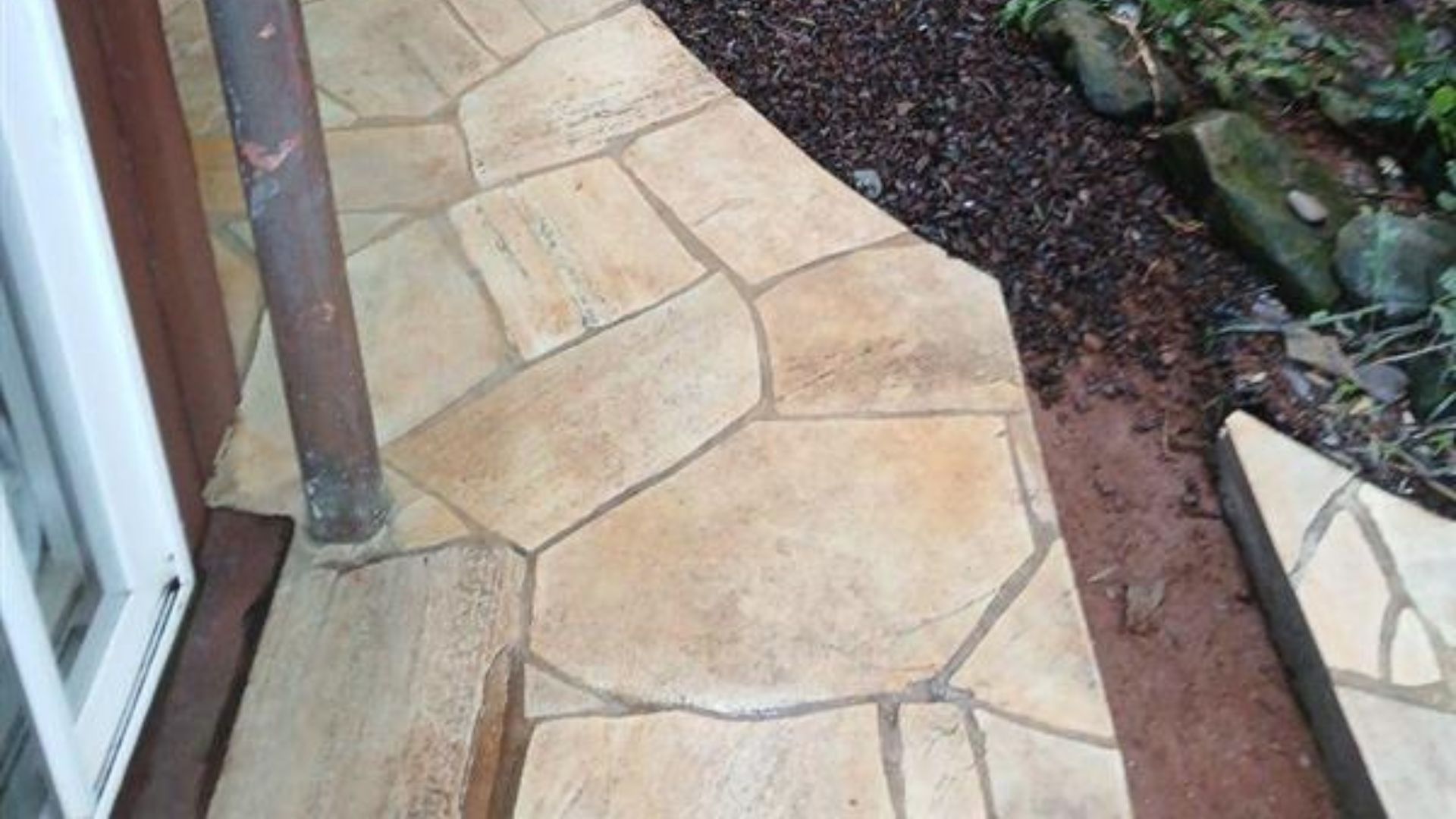 Stone patio walkway beside a house, bordered by soil and rocks with a metal post.