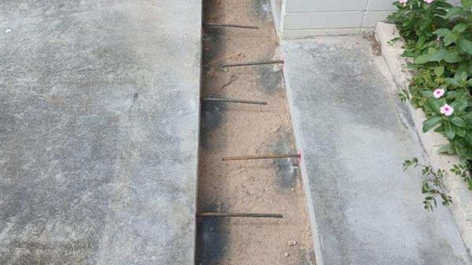 Concrete walkway with a narrow drainage gap and metal bars beside a flower bed