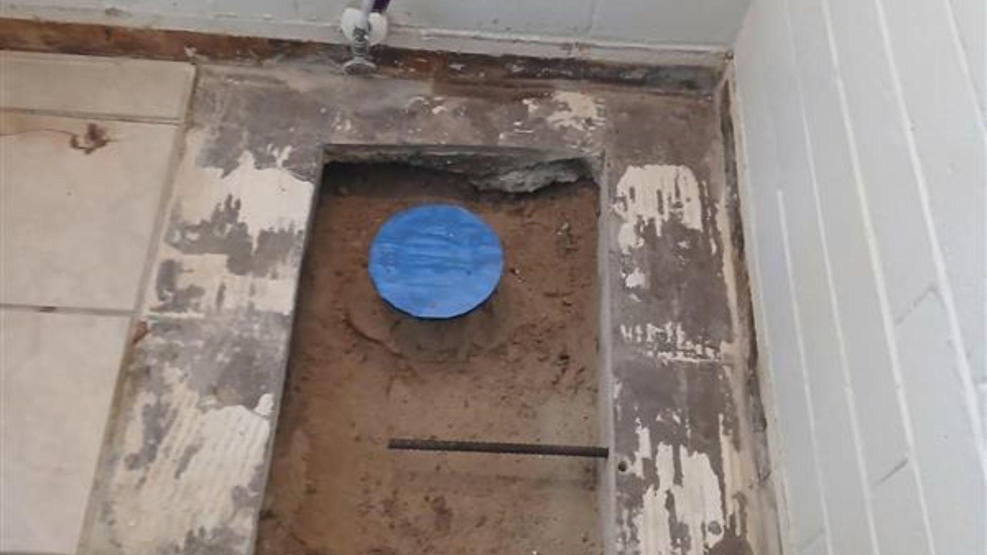Open floor cutout with a blue-capped pipe inside, surrounded by damaged tile and concrete.
