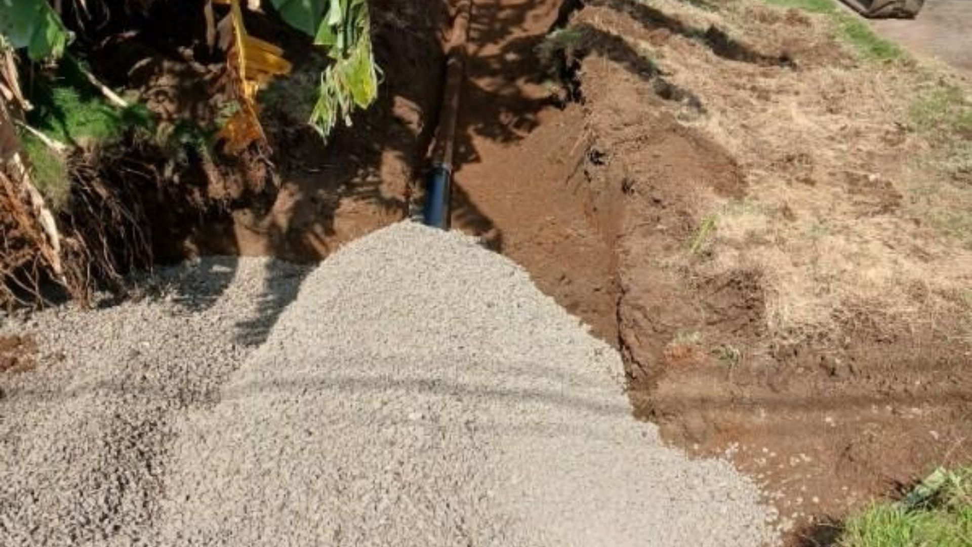 Pile of gray gravel poured into a trench beside an earthen ditch and green vegetation