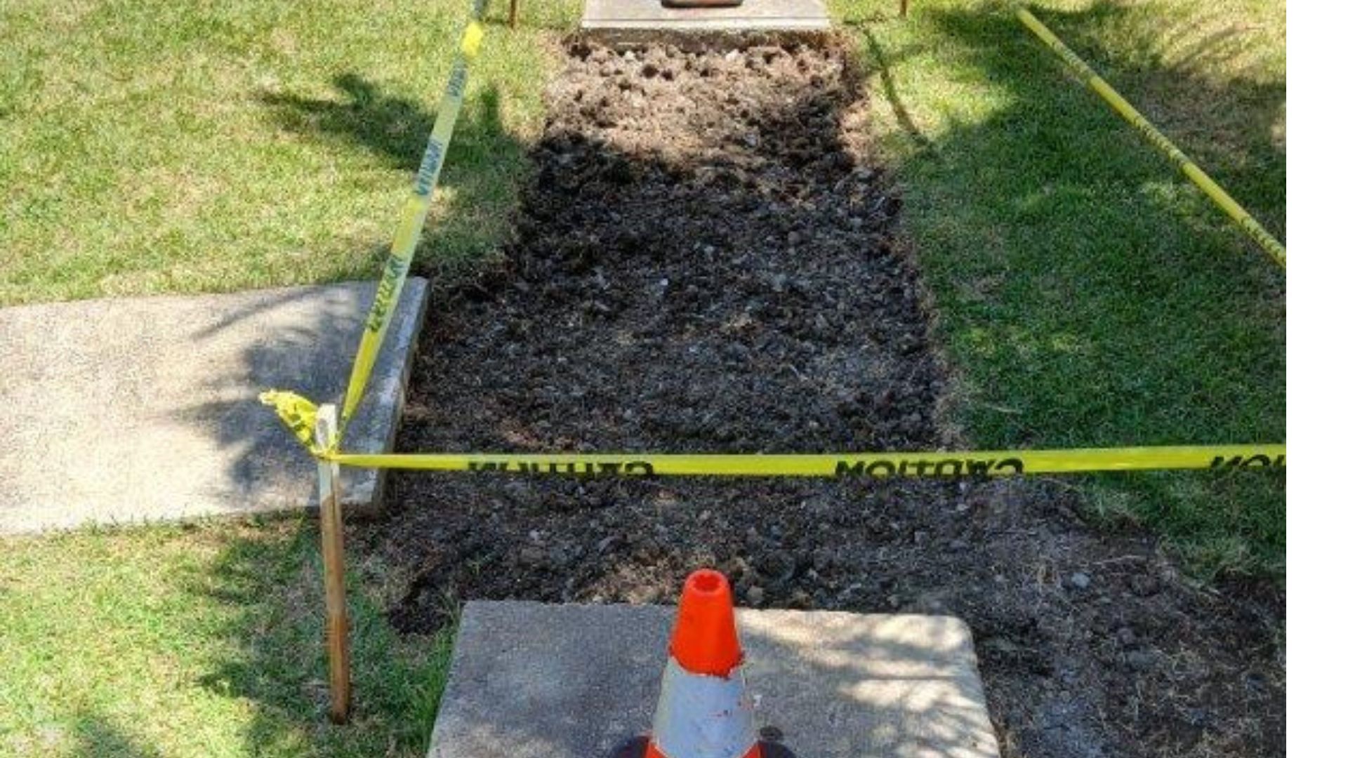 Caution tape blocks a dug-up walkway with gravel, bordered by grass and a traffic cone.