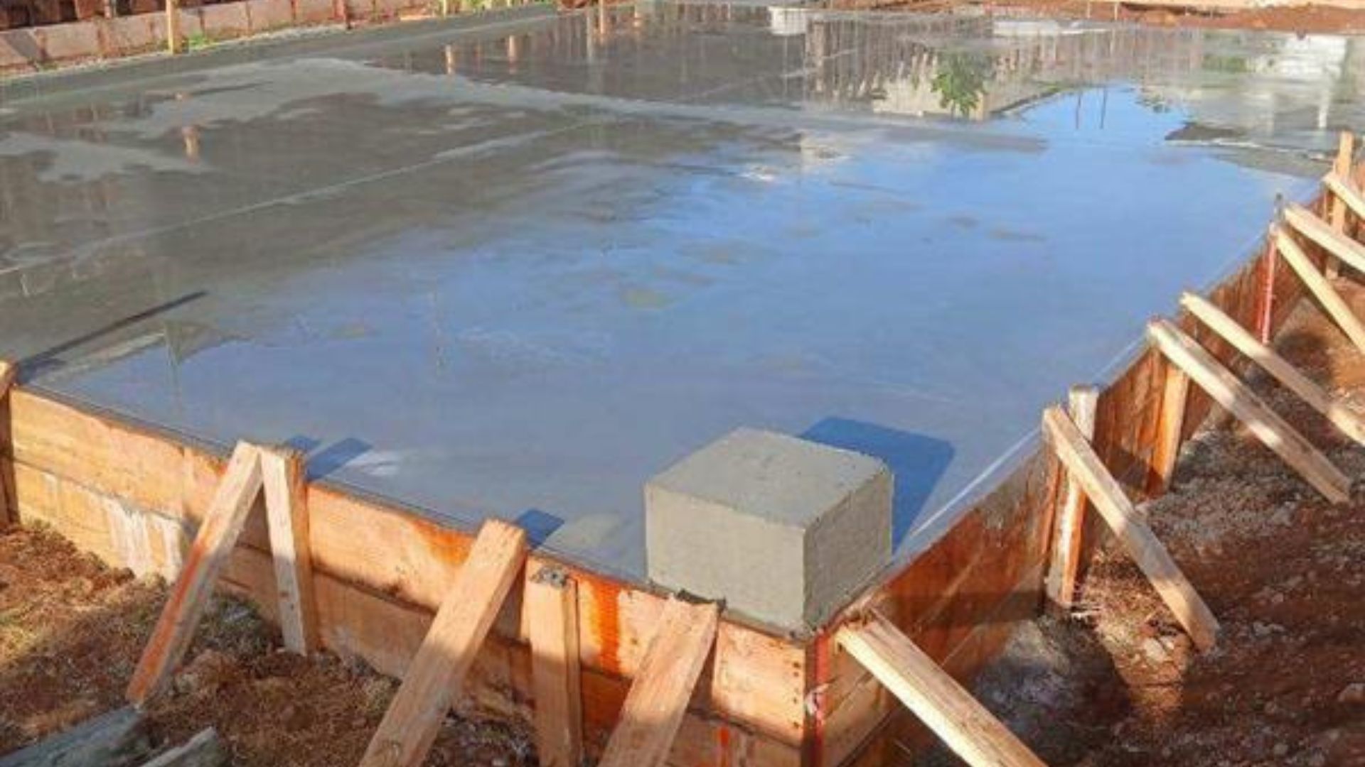 Concrete foundation slab under construction, surrounded by wooden forms and wet surface reflections