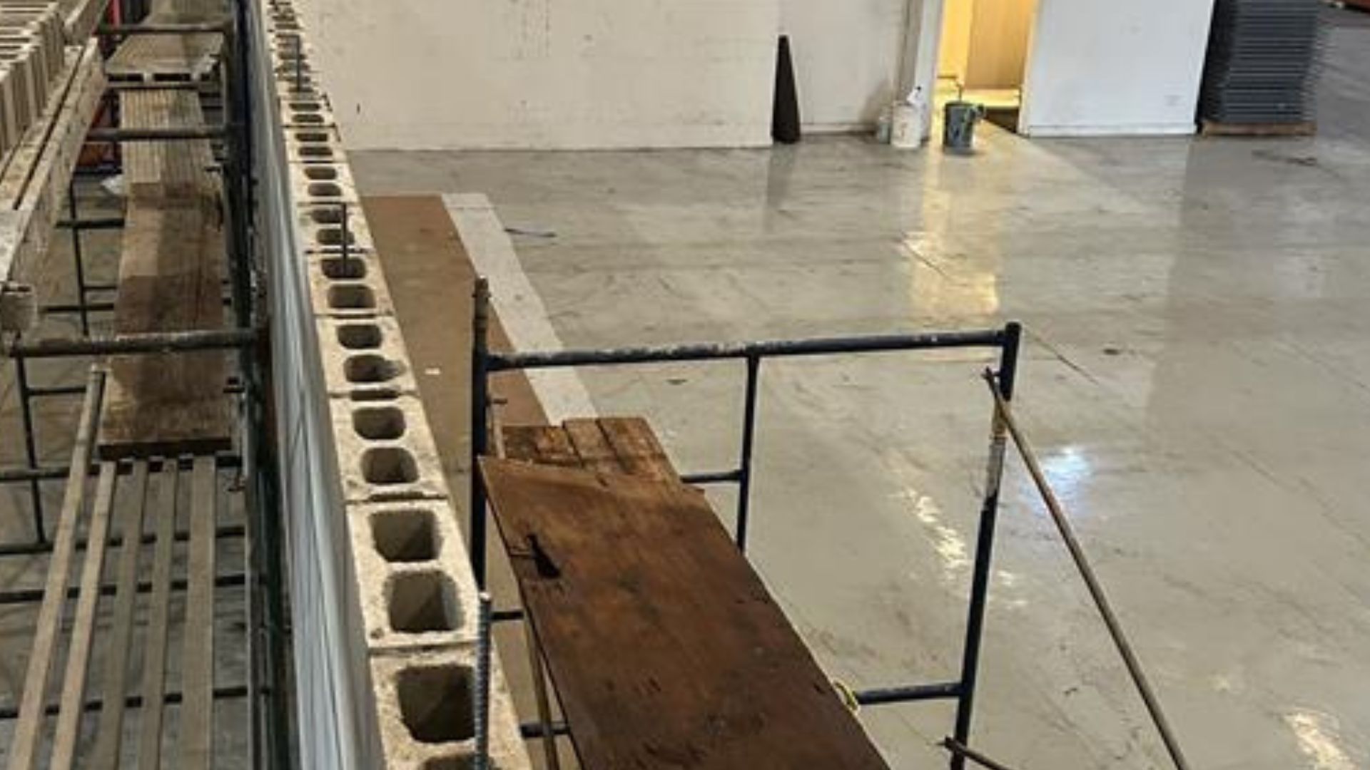 Industrial workspace with concrete floor, cinder blocks, and a wooden plank over metal scaffolding.