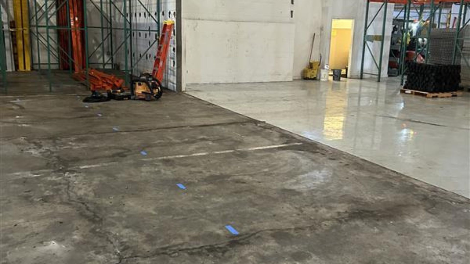 Empty industrial warehouse floor with concrete slabs, construction equipment, and metal shelving.