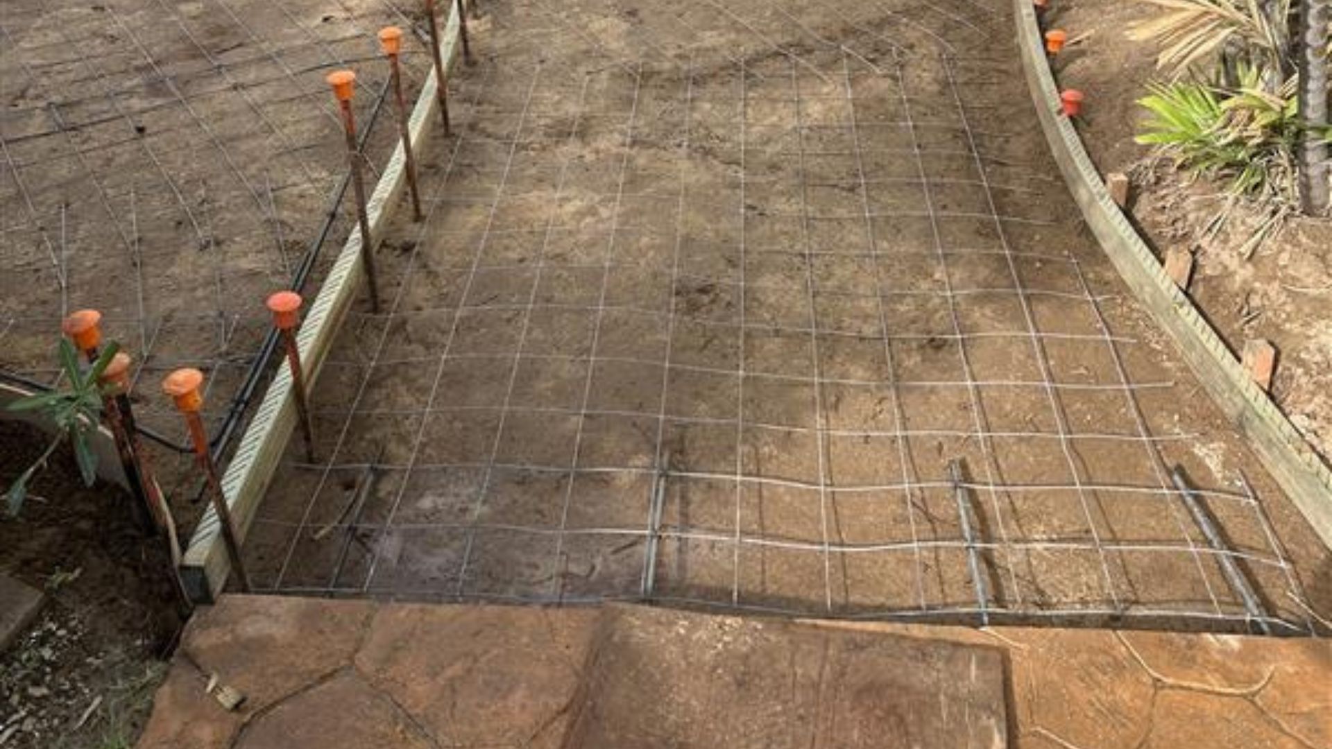 Sloped dirt walkway under construction with wire mesh, bordered by metal posts and orange caps