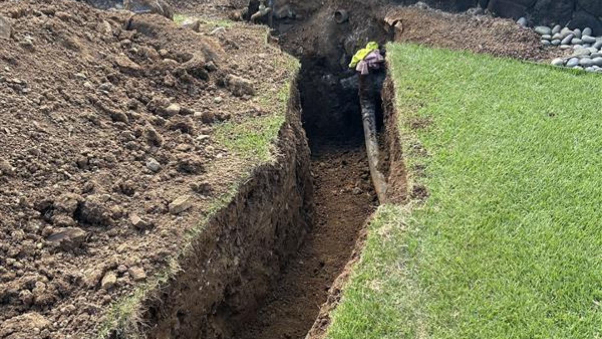 Narrow trench dug beside a green lawn, with an exposed wooden post at the edge