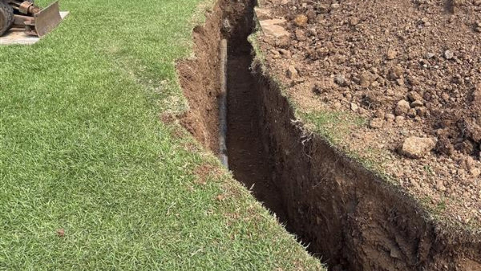 Deep trench cuts beside a grassy lawn, with exposed soil and a dirt pile on the other side.