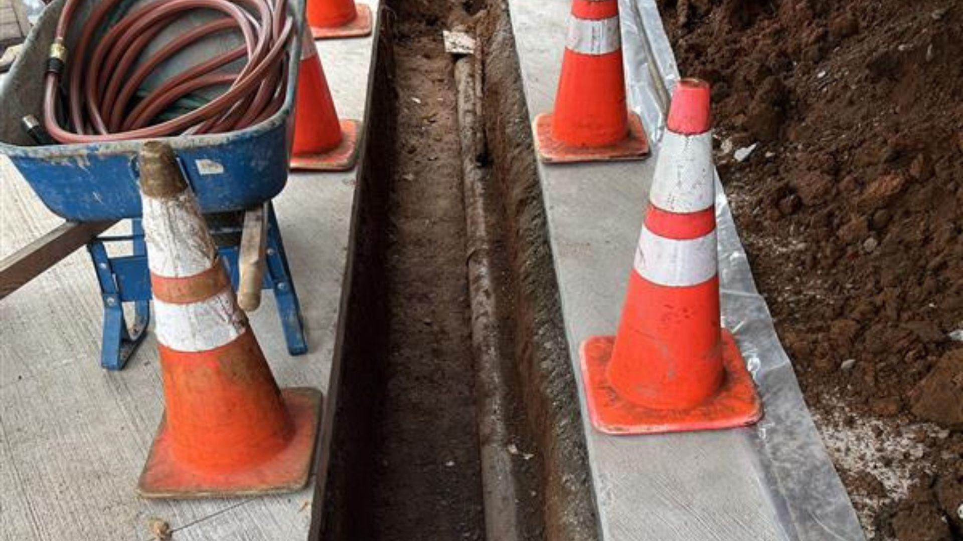 Construction trench beside orange traffic cones and coiled hose on a worksite