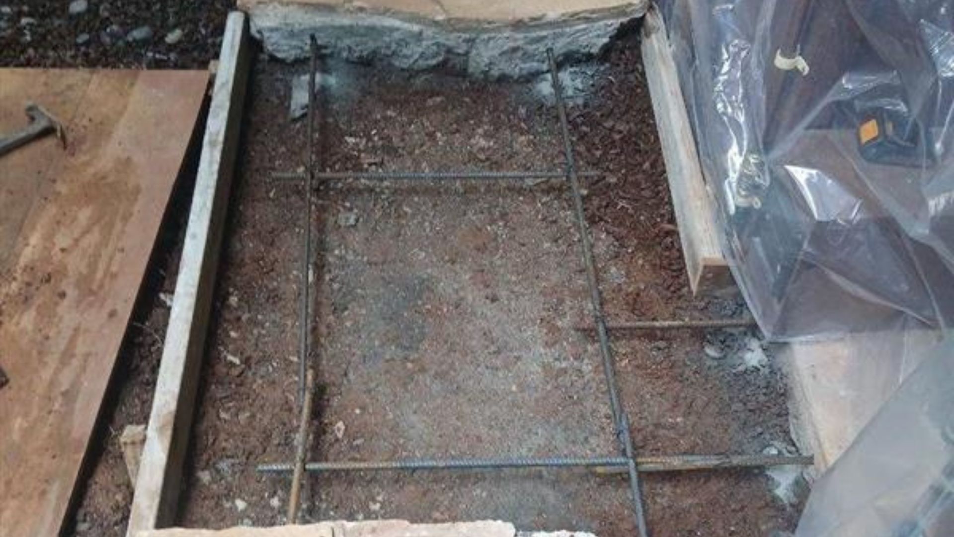 Damaged floor opening with exposed rebar and concrete debris beside plastic-covered construction materials.