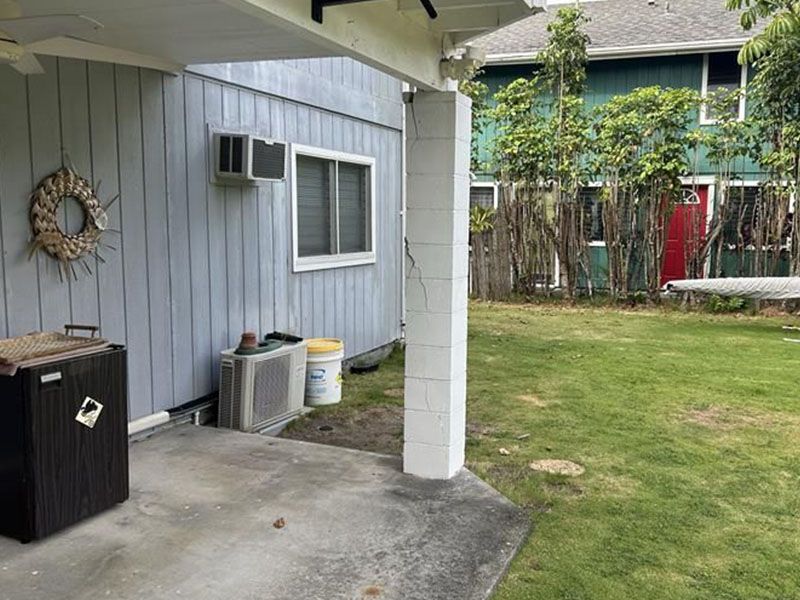 Covered patio beside grassy yard and apartment building, with trash bin, window AC units, and white support post