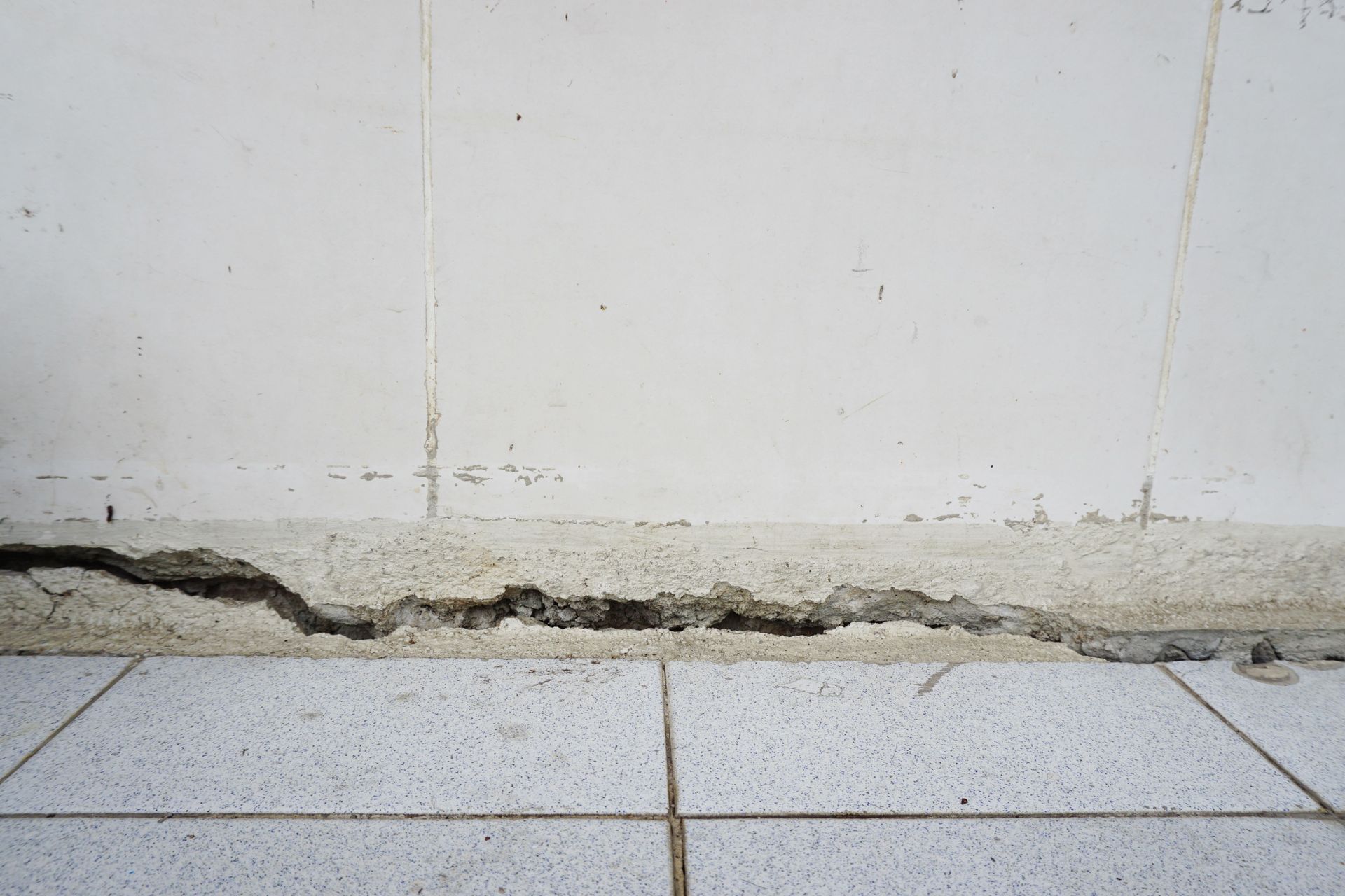 A horizontal crack in the base of a white wall where it meets light-colored tiled flooring.