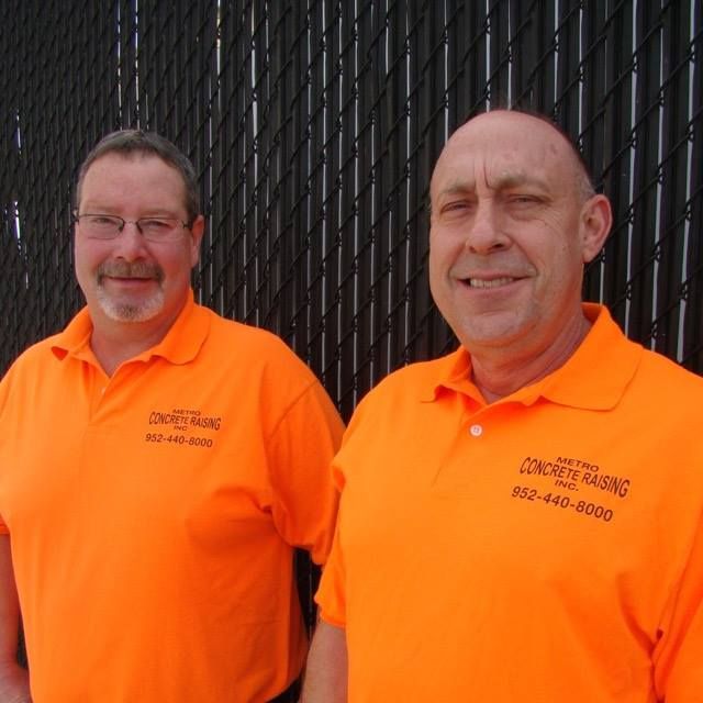 Two people in bright orange work shirts with company logos stand side-by-side in front of a black chain-link fence.