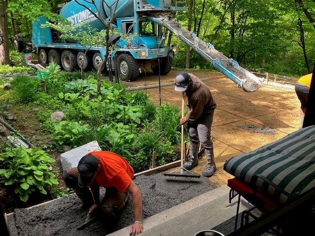 Two construction workers spread wet concrete from a truck chute onto a wooded yard site.