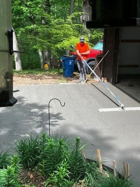 A person in an orange shirt uses a long-handled tool to smooth wet concrete on a driveway near a garage and green trees.