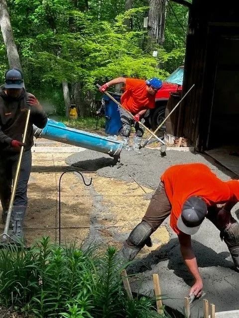 Three construction workers in bright orange shirts and work gear spread wet concrete using tools in a wooded yard.