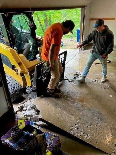 Two people work inside a garage using a skid steer loader to break up and remove a section of damaged concrete flooring.