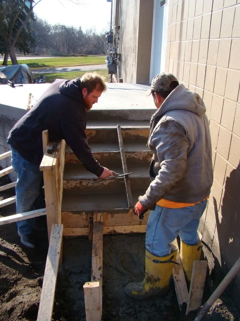 Two workers finish smoothing fresh concrete steps leading to a building entrance using a straightedge tool.