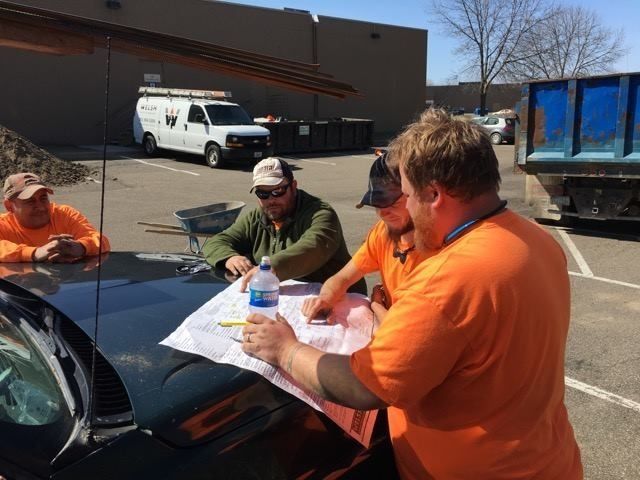 Four construction workers in safety gear review blueprints spread over the hood of a car in a parking lot.