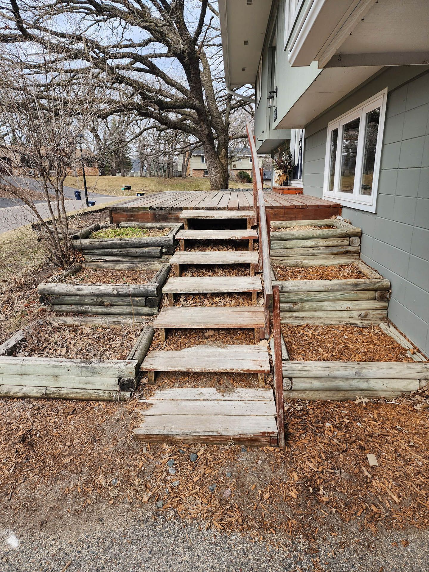A set of weathered wooden steps leading to a deck, flanked by terraced garden beds filled with dry leaves.