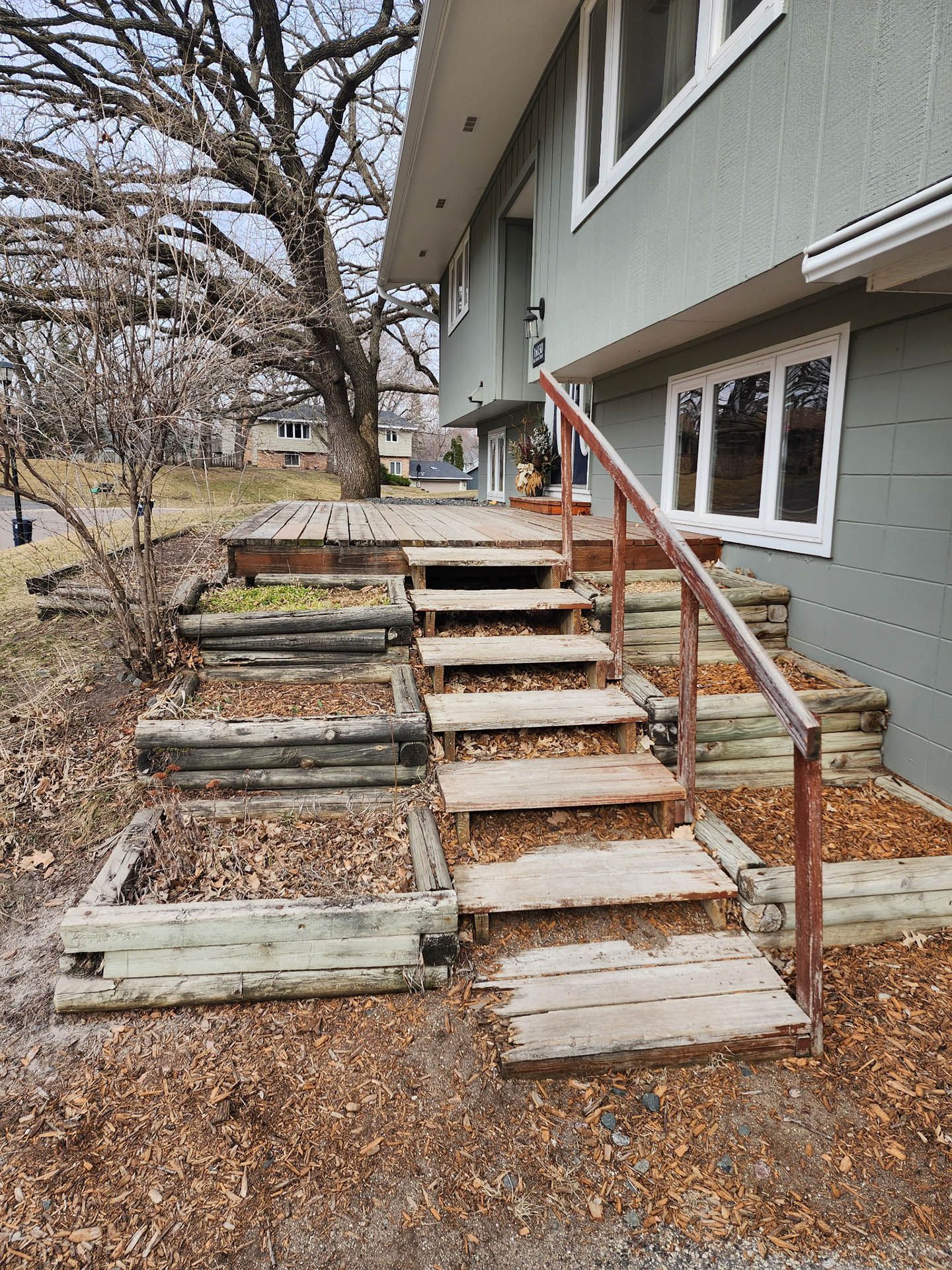 Outdoor wooden steps and terraced planter boxes lead up to a gray house with a wooden deck.