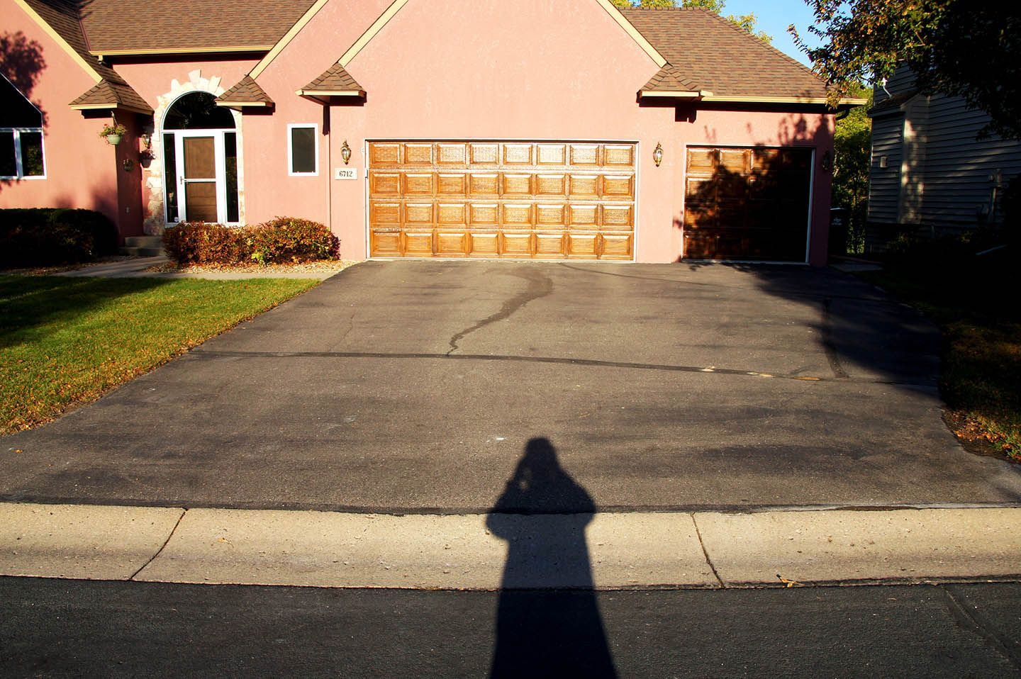 A pink house with a wooden garage door and a cracked asphalt driveway, with a long shadow cast across the foreground.