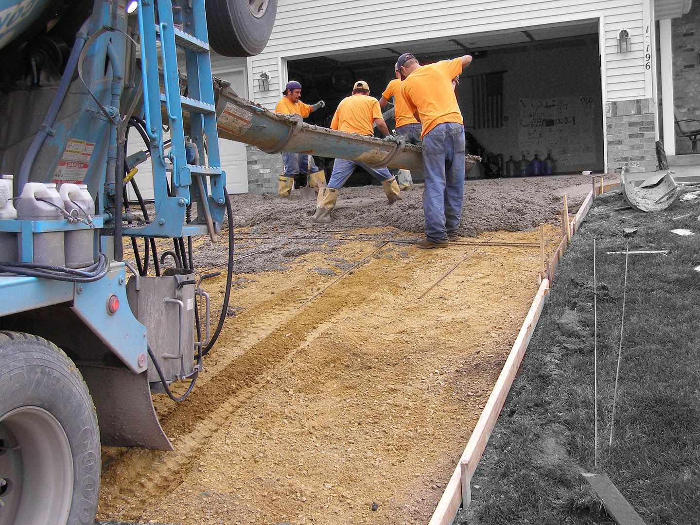 Workers in orange shirts pour wet concrete from a truck mixer onto a prepared driveway base.