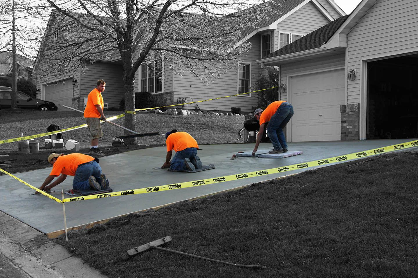 Four people in orange shirts work on a wet concrete driveway in front of a house, enclosed by caution tape.