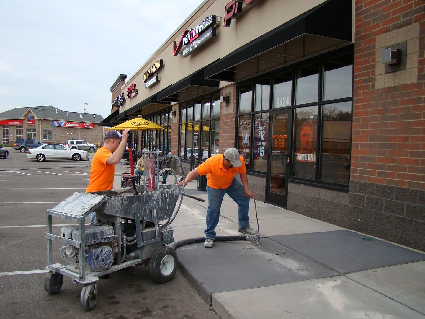 Two workers in bright orange shirts use a mobile concrete sealer machine on a sidewalk outside a retail storefront.