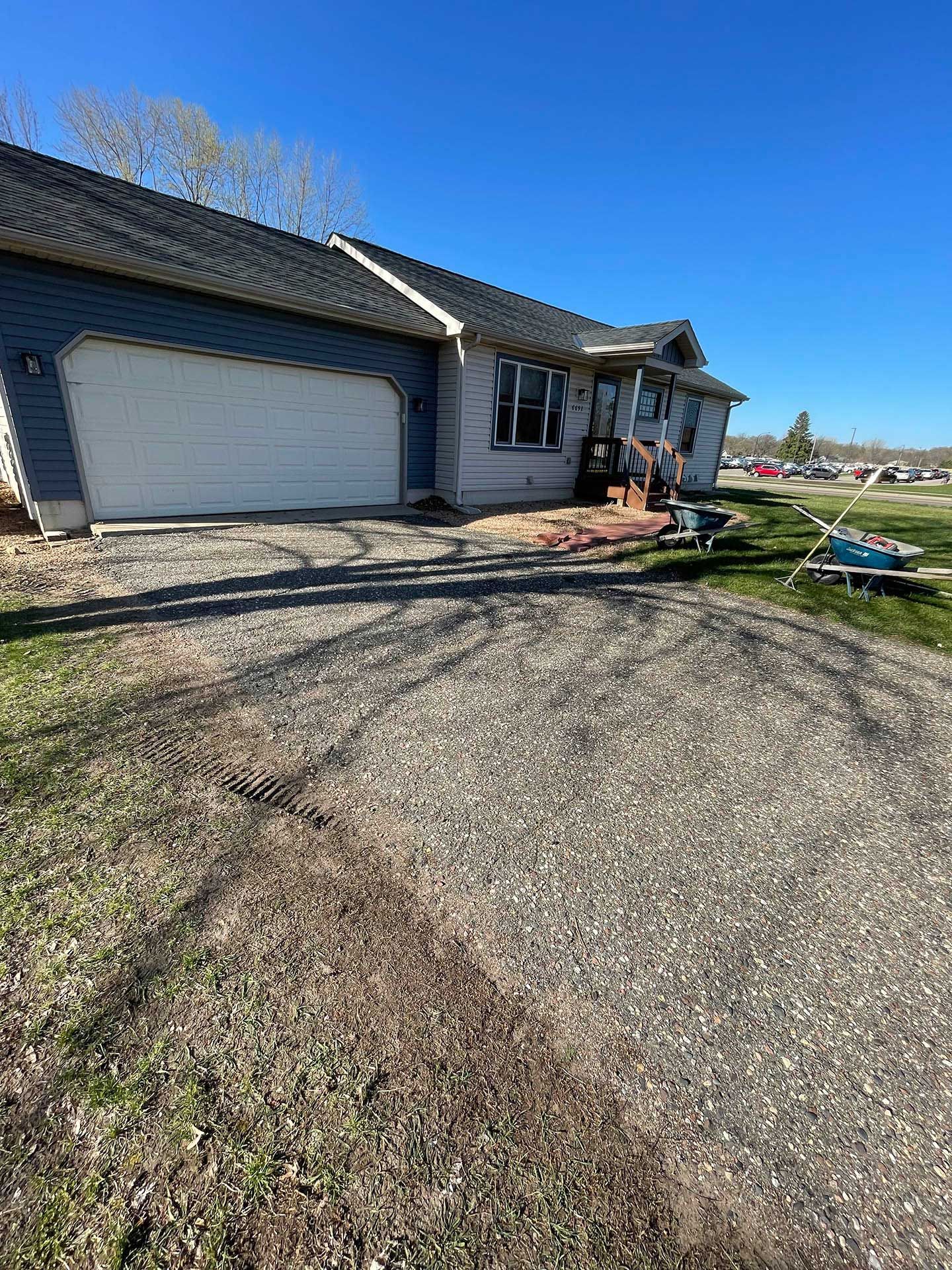 A single-story house with dark siding and a white garage door, set behind a gravel driveway under a clear blue sky.